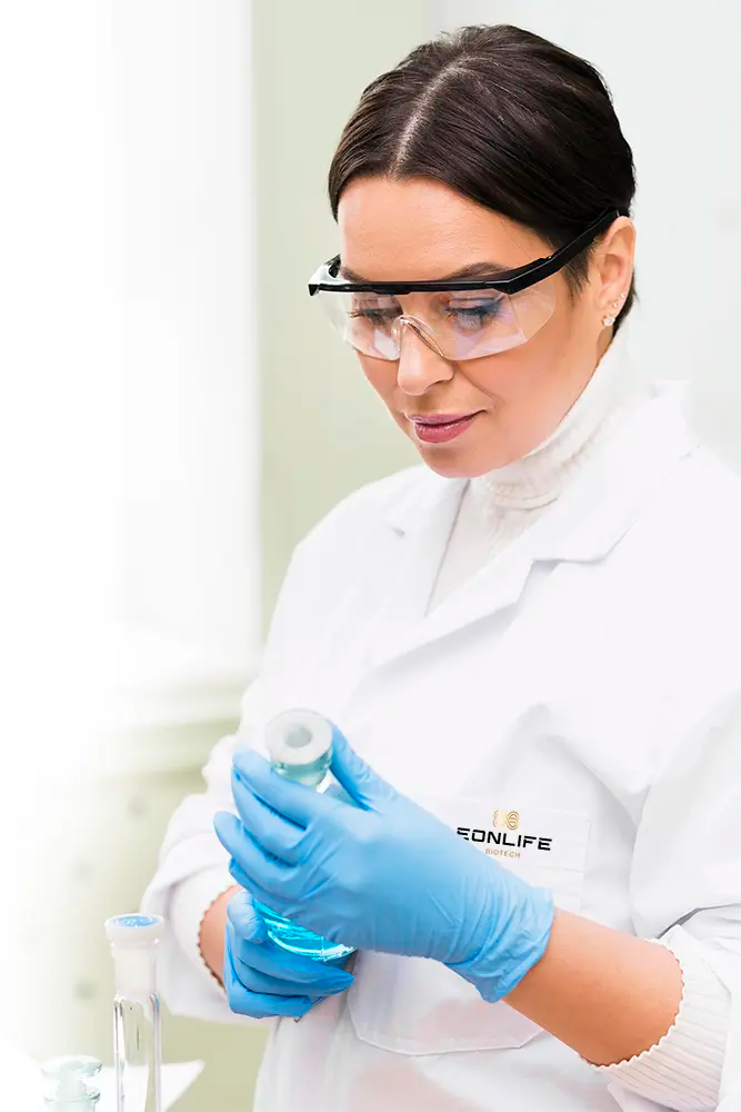Grafika przedstawia kobietę pracującą w laboratorium, ubraną w biały fartuch ochronny z logo EONLIFE Biotech. Ma założone okulary ochronne oraz niebieskie rękawiczki laboratoryjne, a w dłoniach trzyma laboratoryjne naczynie z niebieskim płynem.